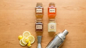 Paper Plane Cocktail Recipe 14 Ingredients for the Paper Plane cocktail including bourbon, Aperol, Amaro Nonino, and fresh lemon juice arranged on a wooden surface.